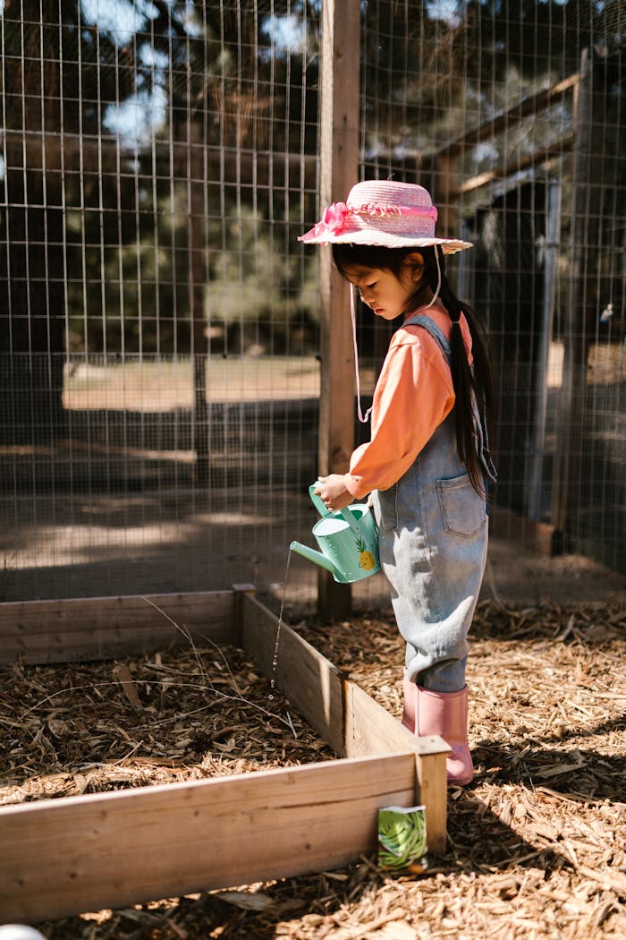 A young girl with a hat watering a garden in a rural setting on a sunny day.